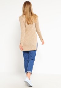 Cardigan long beige en tricot texturé, avec des fentes sur les côtés et un dos ouvert, associé à un jean bleu avec revers et des baskets blanches.