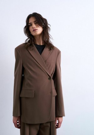 Mujer con blazer marrón extragrande y pantalones a juego, sobre un fondo blanco liso, de pie con postura relajada.