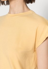 Donna che indossa una maglietta a maniche corte di colore giallo chiaro con scollo rotondo, mostrando collo, spalla e parte superiore del braccio su uno sfondo neutro.