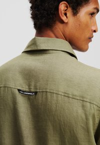 Camicia verde olive in tessuto testo, con colletto classico e etichetta "KARL LAGERFELD" sul retro, vestibilità su misura.