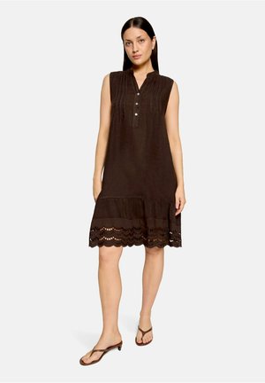 Femme portant une robe sans manches marron foncé longueur genou avec des détails plis et un ourlet festonné, associée à des sandales marron fines sur fond blanc.