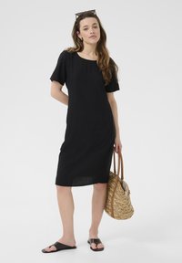 Black linen dress with short sleeves, a round neckline, and a fitted shape. Features a patch pocket and paired with a woven straw bag.