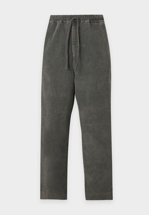 Jeune homme debout portant une chemise à manches longues rayée noir et blanc, un pantalon ample gris foncé et des baskets blanches sur un fond uni.