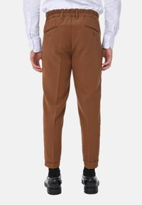 Pantaloni marroni a sigaretta con vita elastica, due tasche posteriori con bottoni e orli a risvolto, indossati con scarpe nere e calzini.