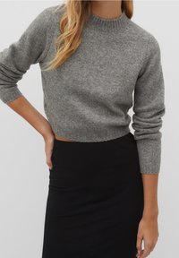 Pull en laine gris avec un col montant et des manches ajustées, associé à une jupe noire. Le pull présente une coupe courte et une texture douce.