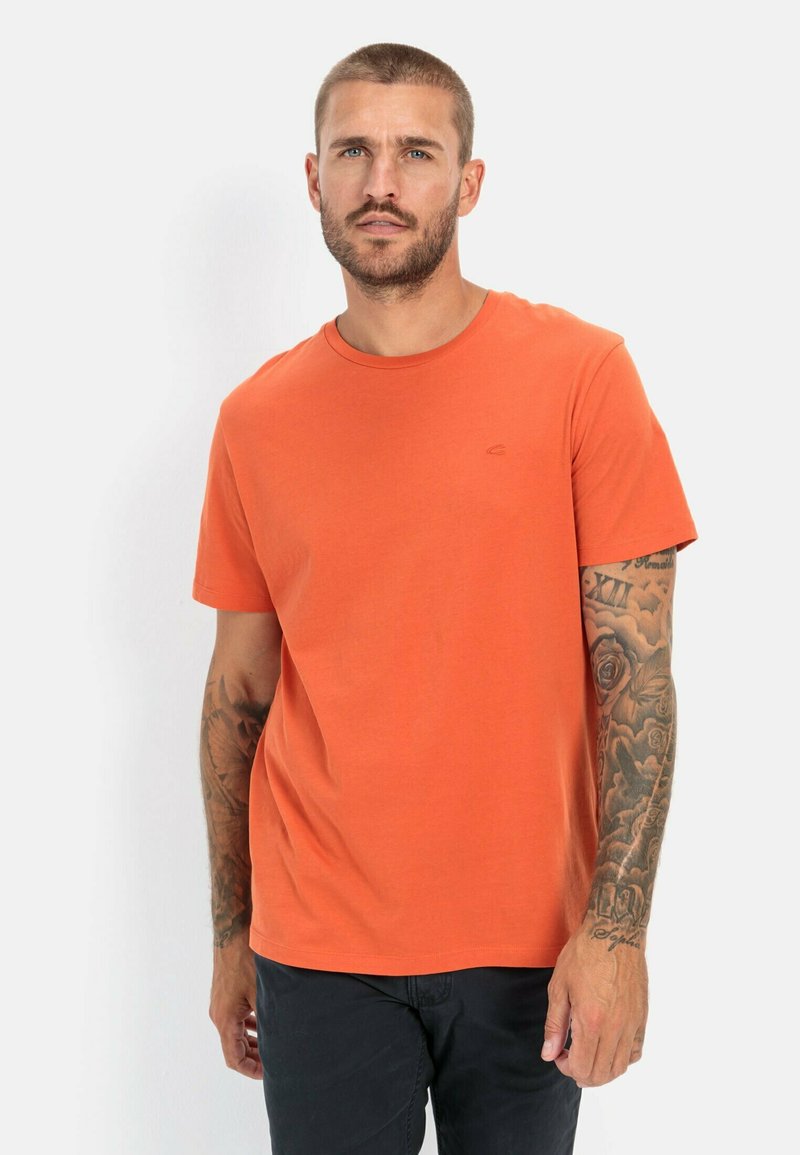 Oranje T-shirt met korte mouwen van katoen en een ronde hals. Bevat subtiele geborduurde logo's. De achtergrond is effen wit, waardoor het shirt opvalt.