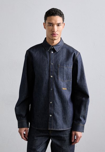 Dark blue denim button-up shirt with a chest pocket, silver snaps, contrasting yellow stitching, and long sleeves. Smooth texture.