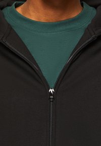 Sudadera negra con cremallera, superpuesta a una camiseta color teal. Textura suave y diseño ajustado visible alrededor del cuello y la cremallera.