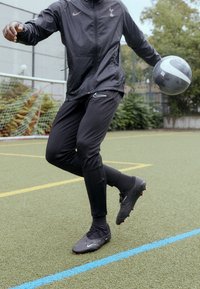 Nike svart träningsoverall med logotyp, åtsittande byxor och fotbollsskor på en konstgräsplan. Håller i en grå och svart fotboll.