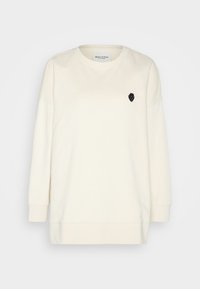 Sweatshirt à manches longues de couleur blanc cassé avec un col rond et un petit emblème noir en forme de fraise sur la poitrine gauche.