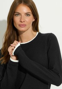 Schwarzer Pullover mit rundem Ausschnitt und weißen Bündchen. Der Stoff ist weich mit einer glatten Textur. Die Ärmel sind leicht lang und bedecken die Handgelenke.
