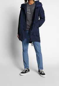 Marinblå parka-jacka med huva, flera fickor och dragsko i nederkant, bärs över en grå sweatshirt och matchas med ljusblå jeans och svarta sneakers.