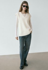 Femme portant des lunettes de soleil noires, un pull sans manches crème par-dessus un haut à manches longues, un jean droit foncé et des chaussures noires, debout sur un sol beige.