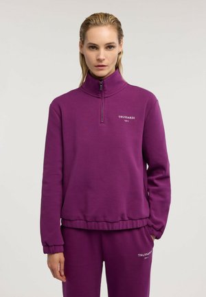 Kobieta o blond włosach ubrana w fioletową bluzę Trussardi na zamek i pasujące spodnie na jednolitym jasnym tle.