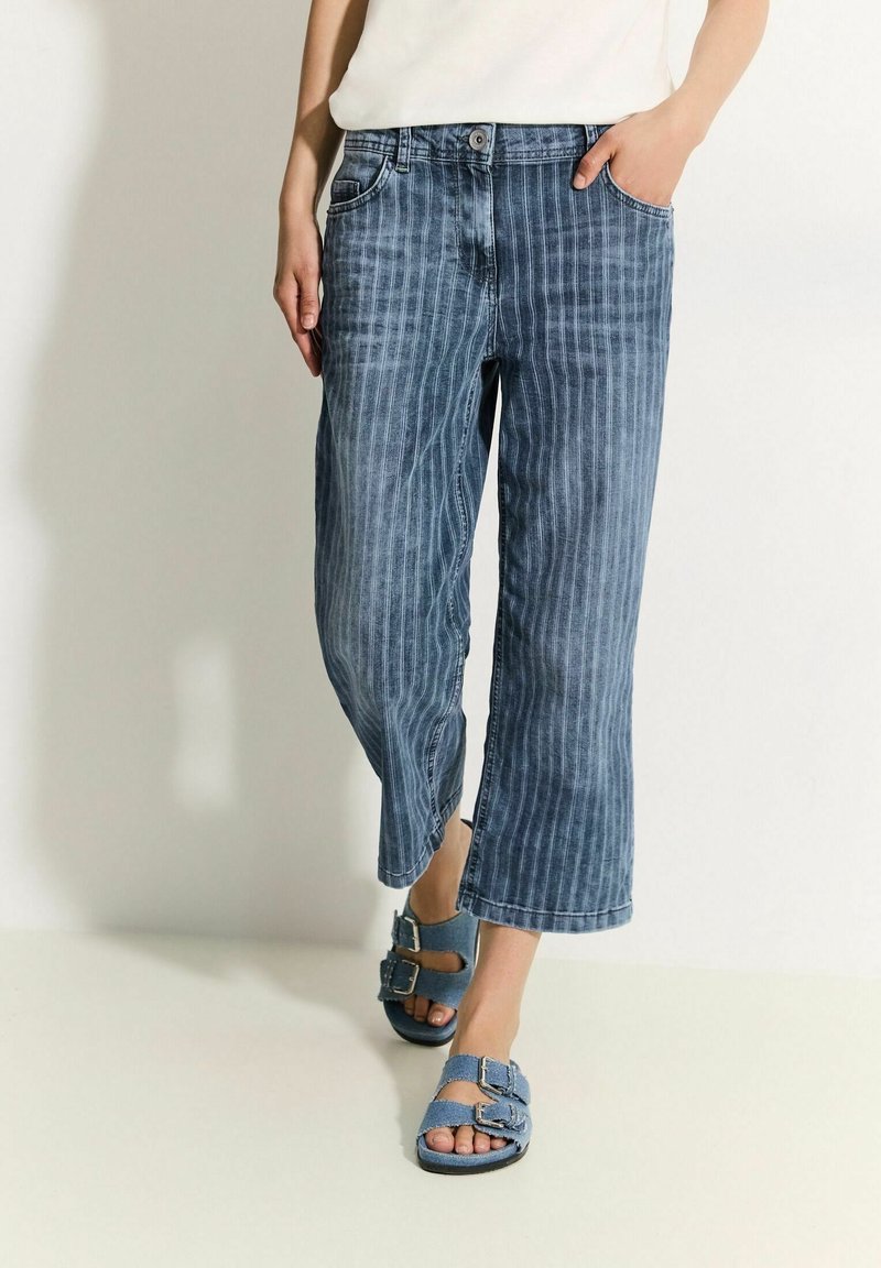 Pantalones vaqueros de pierna ancha con rayas azules, cintura de altura media, bolsillos delanteros y cierre de botón, combinados con sandalias azules de punta abierta.