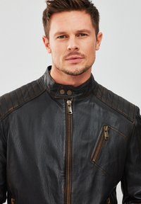 Chaqueta de cuero negra con detalles acolchados en los hombros, cuello alto, cierre delantero de cremallera y bolsillo con cremallera diagonal, con acentos en marrón contrastante.