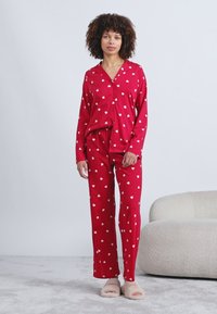 Κόκκινη πιτζάμα που περιλαμβάνει ένα πουκάμισο με κουμπιά και μακριά μανίκια, διακοσμημένο με λευκά σχέδια καρδιάς, σε συνδυασμό με ταιριαστά παντελόνια. Μαλακή υφή, άνετη εφαρμογή.
