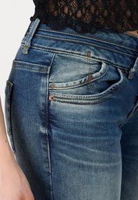 Jeans de mezclilla azul con un diseño ajustado, que presentan un leve desgaste, costuras visibles y detalles metálicos en color cobre cerca de los bolsillos.