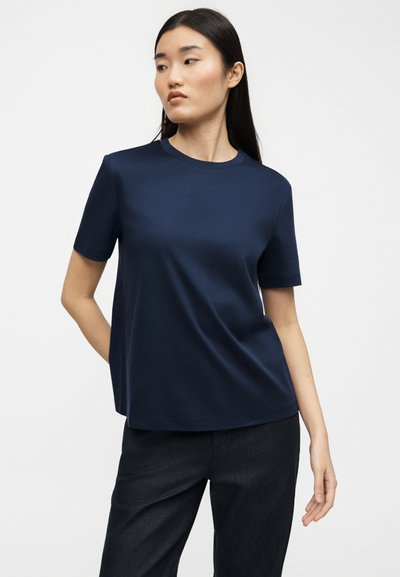 Γυναίκα με μακριά σκούρα μαλλιά, φοράει μπλε κοντομάνικο πουκάμισο και μαύρο παντελόνι, στέκεται με το ένα χέρι πίσω από την πλάτη της.