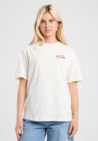 Cream-colored cotton t-shirt with short sleeves, featuring red text and graphic on the left chest. Relaxed fit, soft texture.