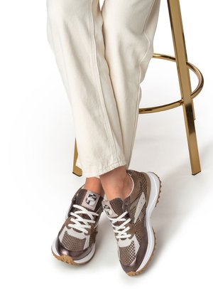 Brown mesh and leather sneakers feature white laces, a patterned design, and a rubber sole, paired with light cream pants.