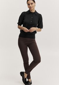 Maglione polo nero lavorato a maglia con maniche corte a sbuffo e colletto con tre bottoni, abbinato a pantaloni marroni aderenti e mocassini neri.