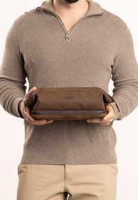 Pochette in pelle marrone con chiusura a zip, caratterizzata da una texture liscia e un logo del marchio sul fronte. Tenuta da un individuo.