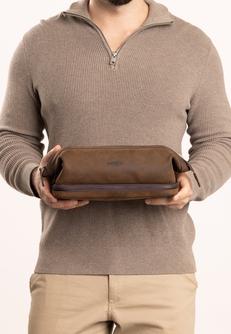 Pochette in pelle marrone con chiusura a zip, caratterizzata da una texture liscia e un logo del marchio sul fronte. Tenuta da un individuo.