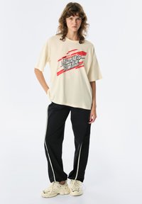 Beige oversized t-shirt met grafische print in rood en zwart; gecombineerd met zwarte trainingsbroek met witte zijstrepen en beige sneakers.