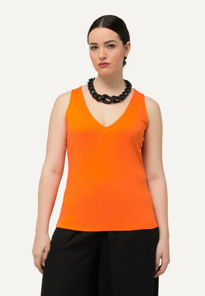 Orange ärmlös topp med V-ringning, tillverkad av ett glatt material. Kombinerad med ett kraftigt svart kedjebyggda halsband. Svarta vida byxor.