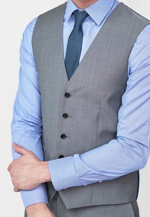 Homme portant un gilet gris boutonné sur une chemise bleu clair avec une cravate bleue à motifs, ajustant son poignet gauche avec sa main droite.