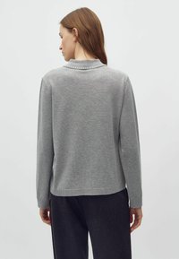 Nő világosszürke hosszú ujjú pulóverben, melynek gallérján gyöngy díszítés látható, háttal áll, sötét nadrággal, egyszerű háttér előtt.