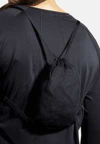 Personne portant un sac à dos noir texturé à cordon sur un t-shirt noir à manches longues, vue de dos et au niveau de l’épaule.