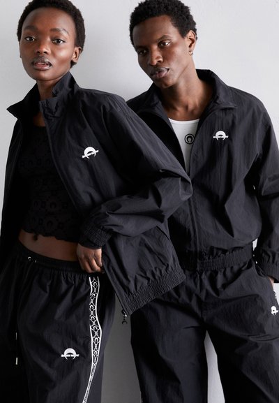 Vêtements de sport noirs, comprenant des vestes et des pantalons en tissu léger, dotés de poignets élastiques et d'accents de logo blancs, présentés ensemble.
