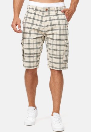 Mann trägt beige karierte Cargo-Shorts mit Taschen und weiße Sneakers, steht mit einer Hand in der Tasche vor schlichtem Hintergrund.