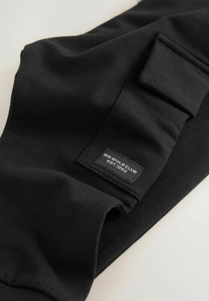 Schwarze Baumwoll-Jogginghose mit geripptem Bündchen, ausgestattet mit einem rechteckigen Patch, auf dem das Markenlabel und das Gründungsjahr seitlich eingestickt sind.