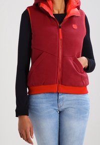 Gilet di pile rosso con cappuccio, accenti arancioni, chiusura frontale con zip e due tasche laterali. Indossato sopra una maglia nera a maniche lunghe.