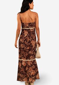 Mujer con un vestido maxi floral marrón de tirantes finos, sosteniendo un bolso beige, de espaldas frente a un fondo blanco liso.