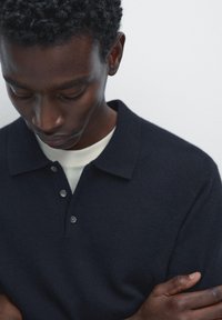 Navy knitted polo shirt with a collar, short sleeves, and three-button placket, layered over a light-colored crew-neck shirt.