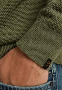 Maglione a maglia verde oliva con motivo testurizzato e polsini a coste. Presenta un piccolo etichetta sul polsino. Mano poggiata su jeans blu scuro.