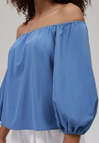 Blå, off-the-shoulder bluse med puffermer, lavet af glat stof. Har samlede detaljer ved halsudskæringen og elastiske manchetter.