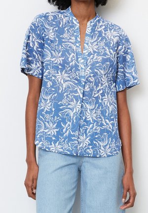 Femme portant un chemisier bleu à manches courtes avec imprimé floral blanc associé à un jean bleu clair sur fond blanc.