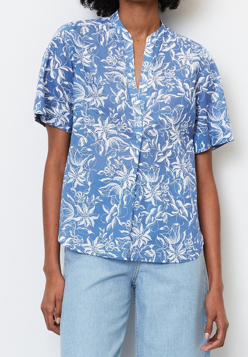 Femme portant un chemisier bleu à manches courtes avec imprimé floral blanc associé à un jean bleu clair sur fond blanc.