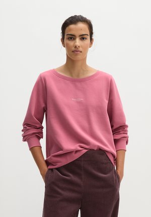 Jeune femme aux cheveux foncés attachés porte un sweat-shirt rose à manches longues et un pantalon en velours côtelé marron foncé, les mains dans les poches.