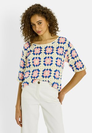 Mujer con pelo rizado que lleva una blusa de manga corta tejida a ganchillo con patrones cuadrados en rosa, azul y crema, y pantalones blancos de tiro alto.