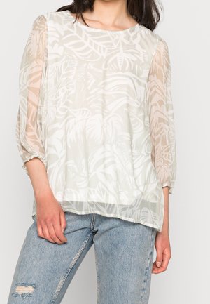 Femme portant un chemisier blanc transparent à manches longues avec des motifs de feuilles et un jean bleu clair déchiré, debout devant un fond uni.