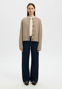 Cardigan en tricot beige avec col rond, fermée par des boutons à l'avant et des poignets côtelés, associé à un jean large bleu foncé et des chaussures brunes.