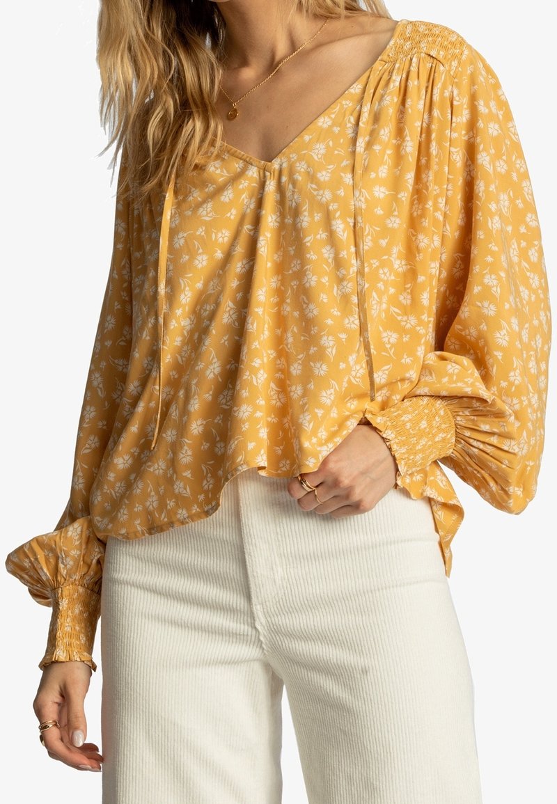 Blouse florale jaune avec un décolleté en V, des manches froncées et un tissu doux et léger ; présente une coupe ample et des poignets élastiques.