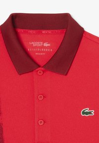 Röd polo skjorta med en strukturerad tyg, med en mörkröd krage, två knappar och en grön krokodil-logga på vänster bröst. Normal passform.