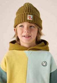 Een jong kind met een geel-groene gebreide muts en een kleurgebloemde hoodie met een glimlachend gezicht patch, kijkend een beetje opzij en glimlachend.